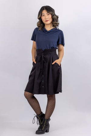 The Natalie skirt, a flared gored skirt pattern for women, with a button front, made in black viscose twill, front view with hands in slash pockets.
