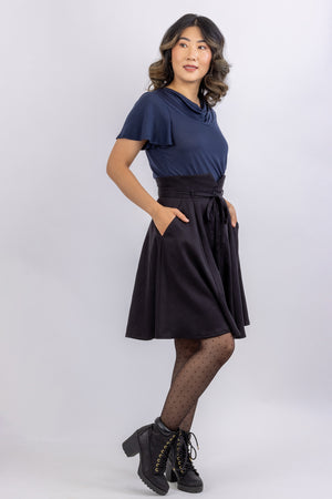 The Natalie skirt, a flared gored skirt pattern for women from Forget-me-not Patterns, with a button front, made in black viscose twill, side view with hands in slash pockets.