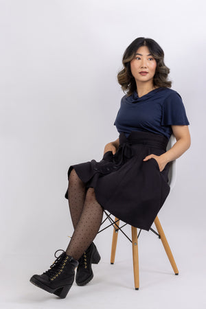 The Natalie skirt, a flared gored skirt pattern for women, with a button front, made in black viscose twill,  seated view with hands in slash pockets.